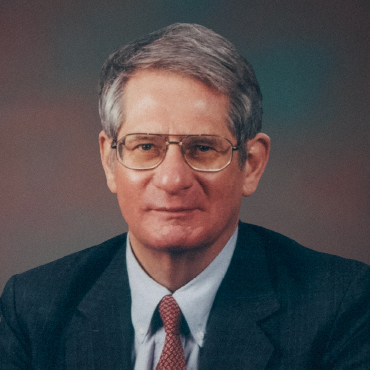 Portrait of Edward Roberts wearing glasses, a dark suit, light blue shirt, and red patterned tie, posed against a softly blurred studio background.