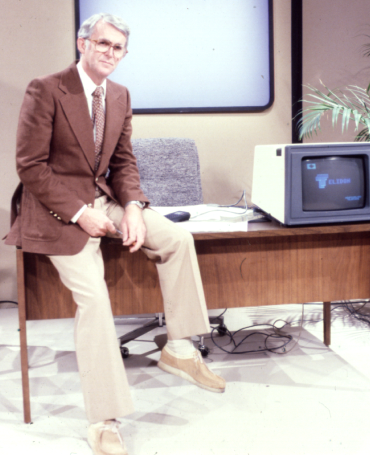 Dr. Max House sits on the edge of his desk next to a monitor with a large screen on the wall behind him