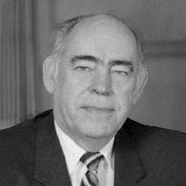 A black and white portrait of Leslie Harris, sitting in a room. He is wearing a suit and has pen and paper Infront of him.
