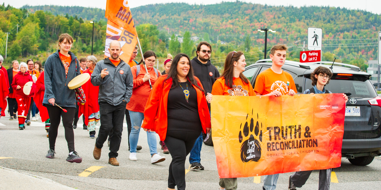 A group of people in orange t-shirts march along a street in Corner Brook with a Truth and Reconciliation banner