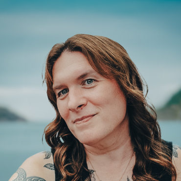 Color portrait of Rhea Rollmann with long wavy brown hair, wearing a sleeveless top, smiling outdoors with water and hills in the blurred background.