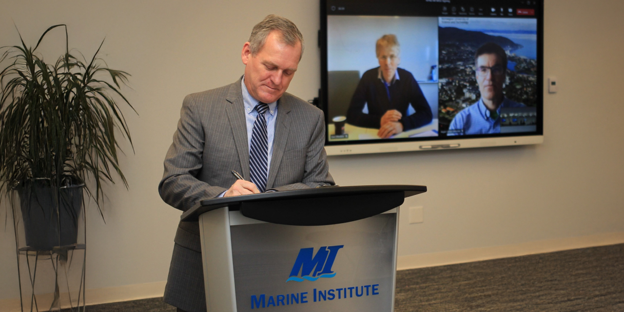 Rob Shea stands at a Marine Institute podium and signs a document while two people look on from a large screen in the background