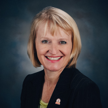 Portrait of Shona Perry-Maidment smiling, wearing a dark blazer with a small lapel pin and a light green top, posed against a dark studio background.
