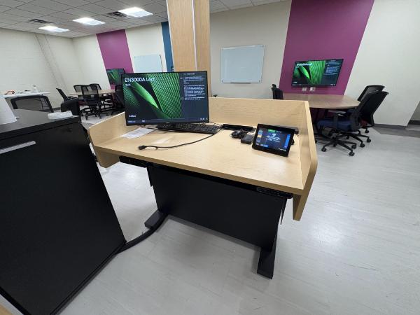 A photo of a classroom, taken from behind a desk with a large monitor and smaller monitor on it. There are long tables with chairs around them and individual monitors for each table.