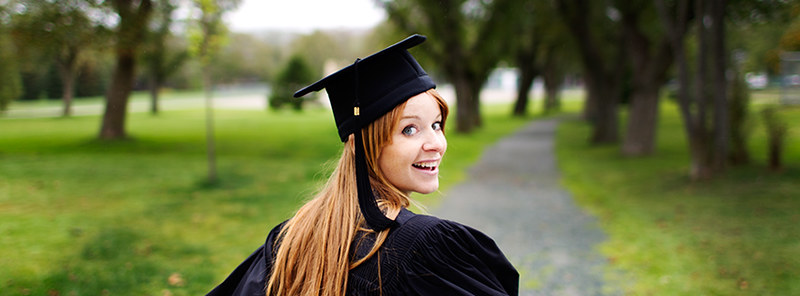A smiling graduating student wearing a cap and gown. There are tree in the background