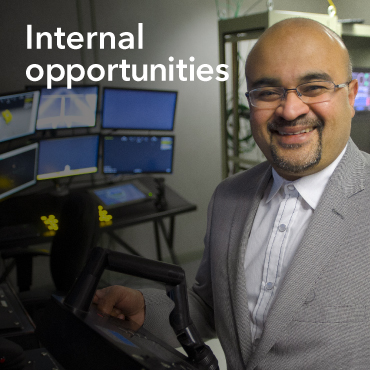 Male employee in a white shirt and grey blazer standing in front of computer screens.  The words Internal Opportunities are on the top left of the image. 