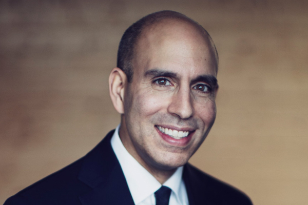 Headshot of Justin Ladha smiling with a black suit with a white shirt and black tie