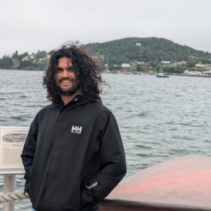 A young man with long brown hair stands in front of the ocean with a coastal community visible in the background