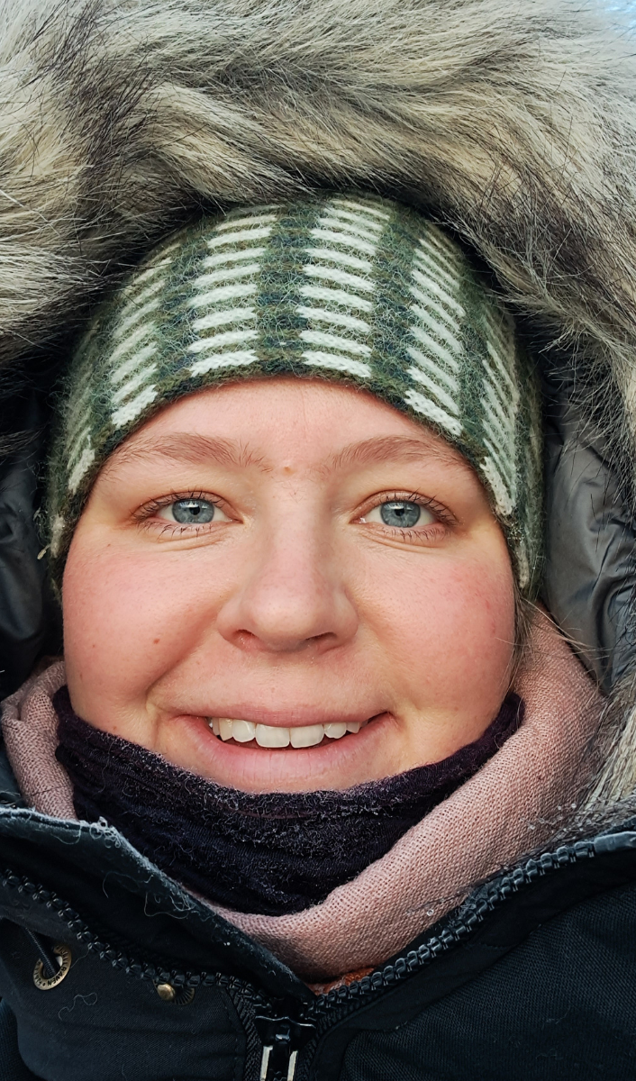 A young blonde woman wearing a pink scarf and a green checkered hat with a fluffy fur hood wrapped around her head