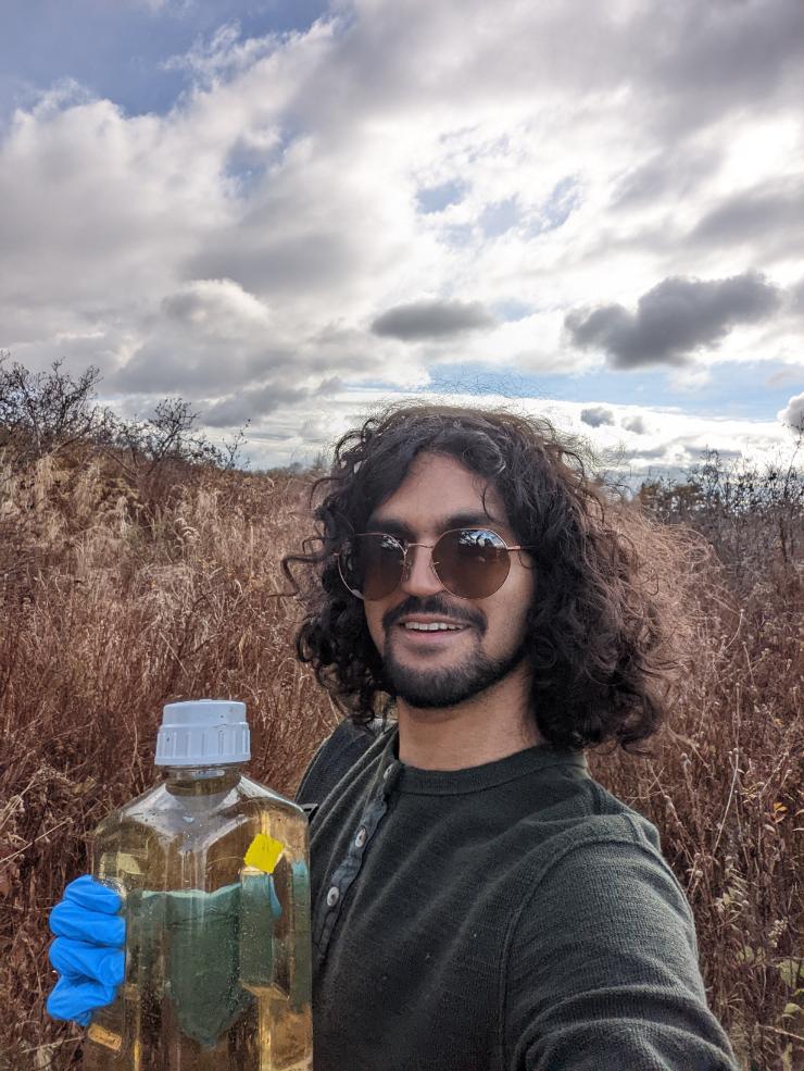 A picture of a young man with long brown hair standing in a field of yellow trees holding a plastic bottle