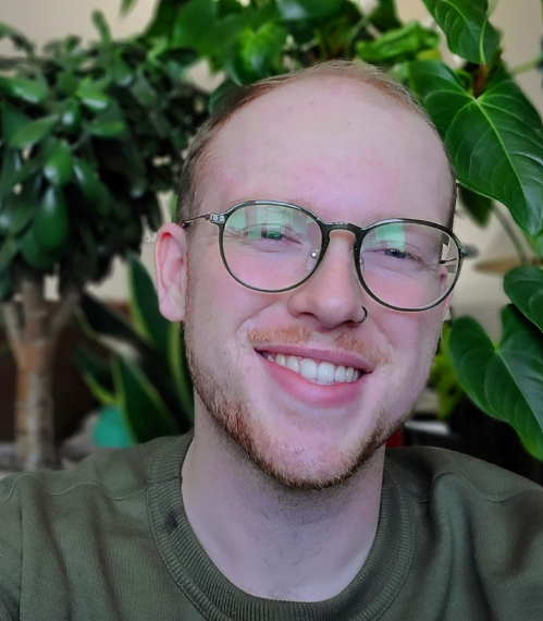 A person with short red hair and glasses with trees in the background