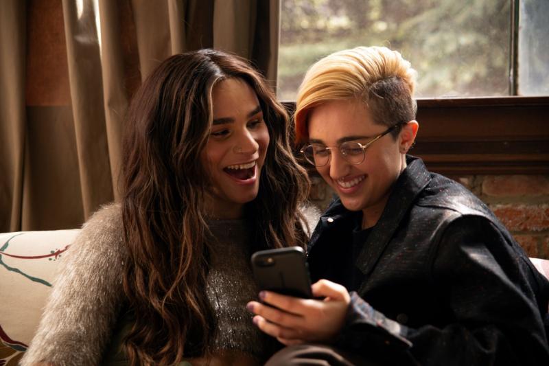 A transfemine non-binary person and transmasculine gender-nonconforming person looking at a phone and laughing